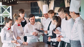 Mehrere Personen in Gastronomiegewändern, wie Schürzen und Kochhauben, stehen in Restaurant und blicken auf weitere Person mit Clipboard und Stift in Händen