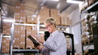 Lächelnde Person mit Brille tippt auf Tablet, im Hintergrund verschwommen Regale mit gestapelten Kartons
